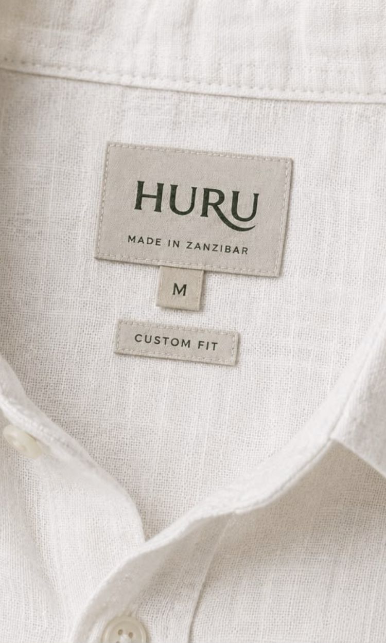 Woven HURU Made in Zanzibar label inside the collar of a white linen shirt, above a size M tab